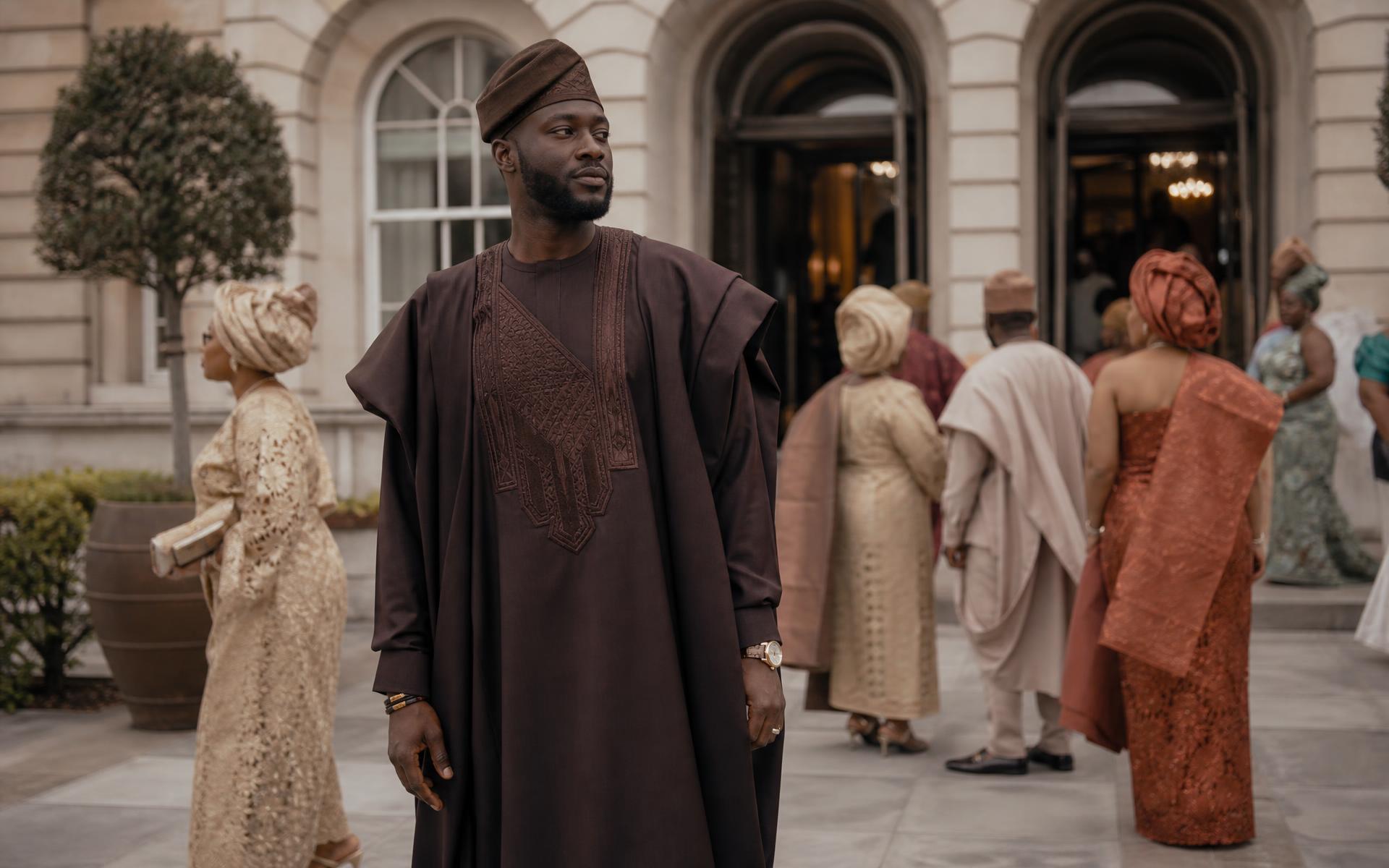 An aubergine agbada worn arriving at a London wedding among guests in aso-ebi