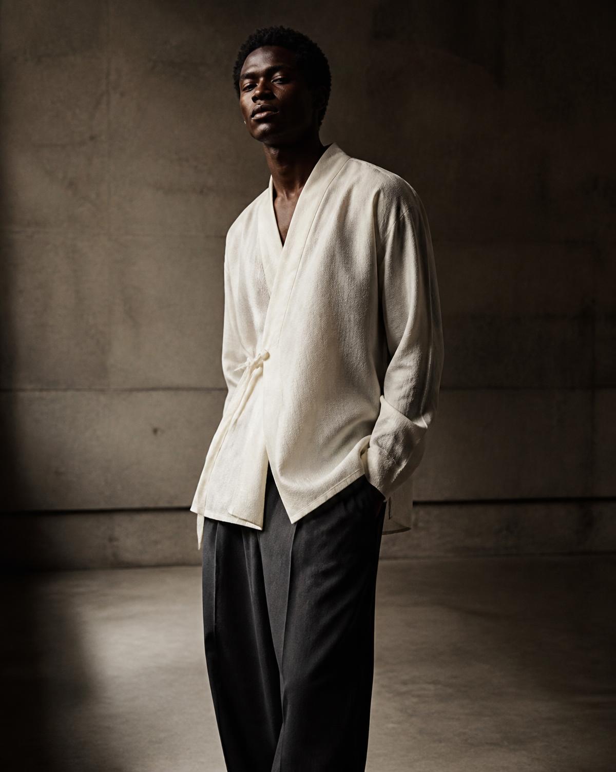 An unstructured ivory jacket worn open over a black trouser in a dim interior